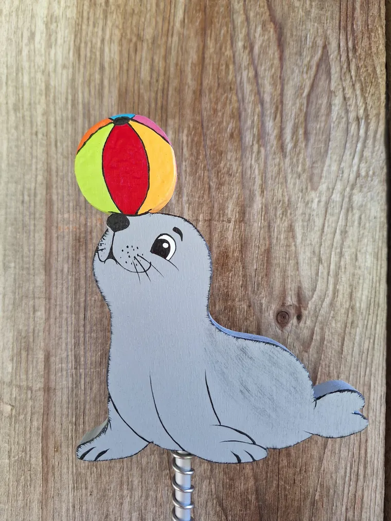 Handbemalte Robbe mit buntem Ball als Gartenstecker vor einer Holzwand.