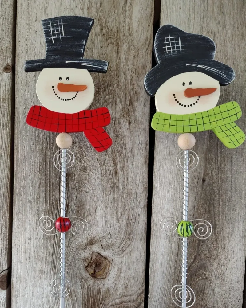 Zwei handbemalte Schneemann-Gartenstecker mit rotem und grünem Schal vor einer Holzwand.